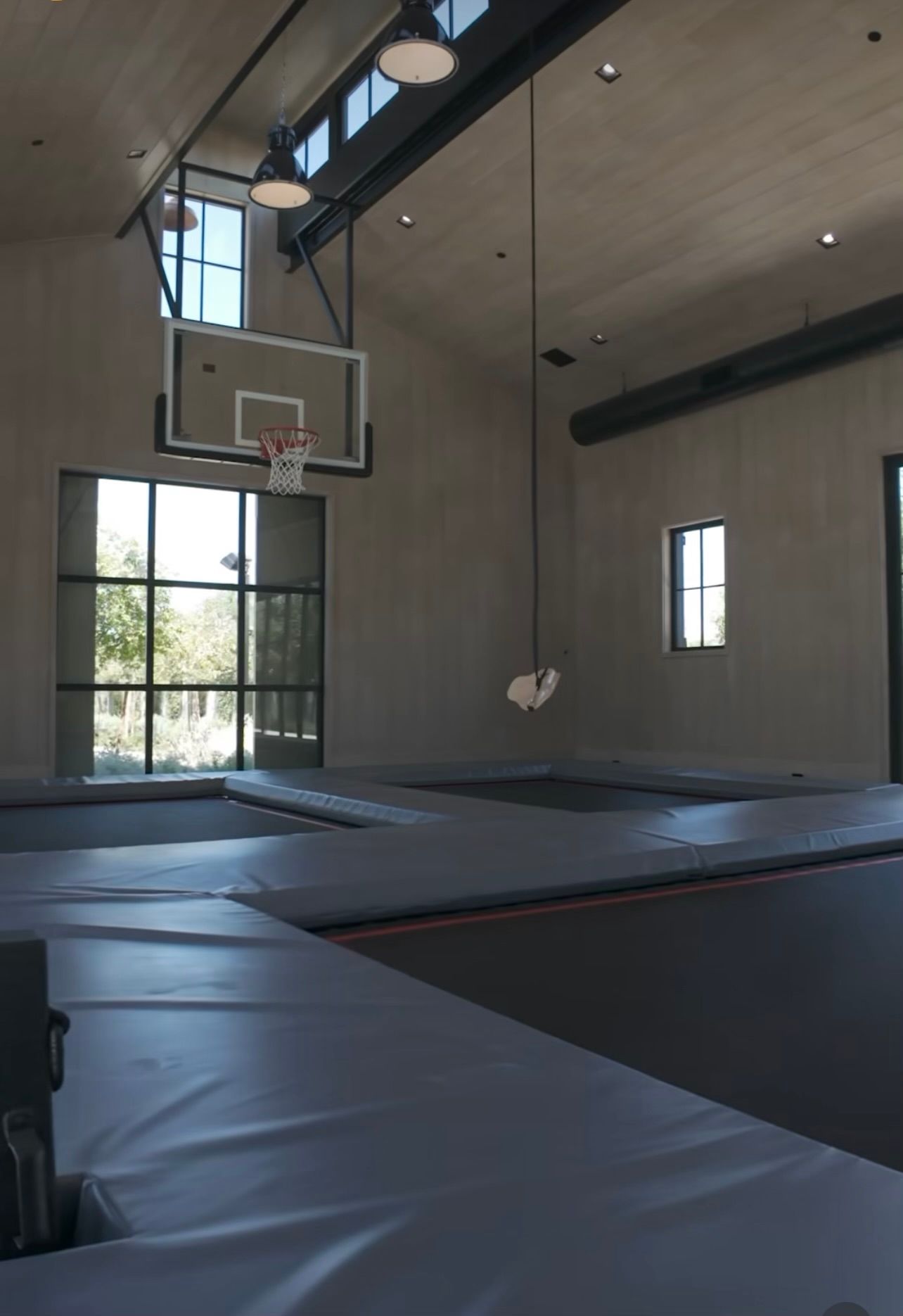 Indoor gym with trampoline courts and a basketball hoop beneath tall windows and wood ceiling panels