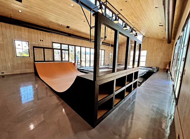 Indoor skate park with a wooden half-pipe and ramps inside a bright, unfinished room