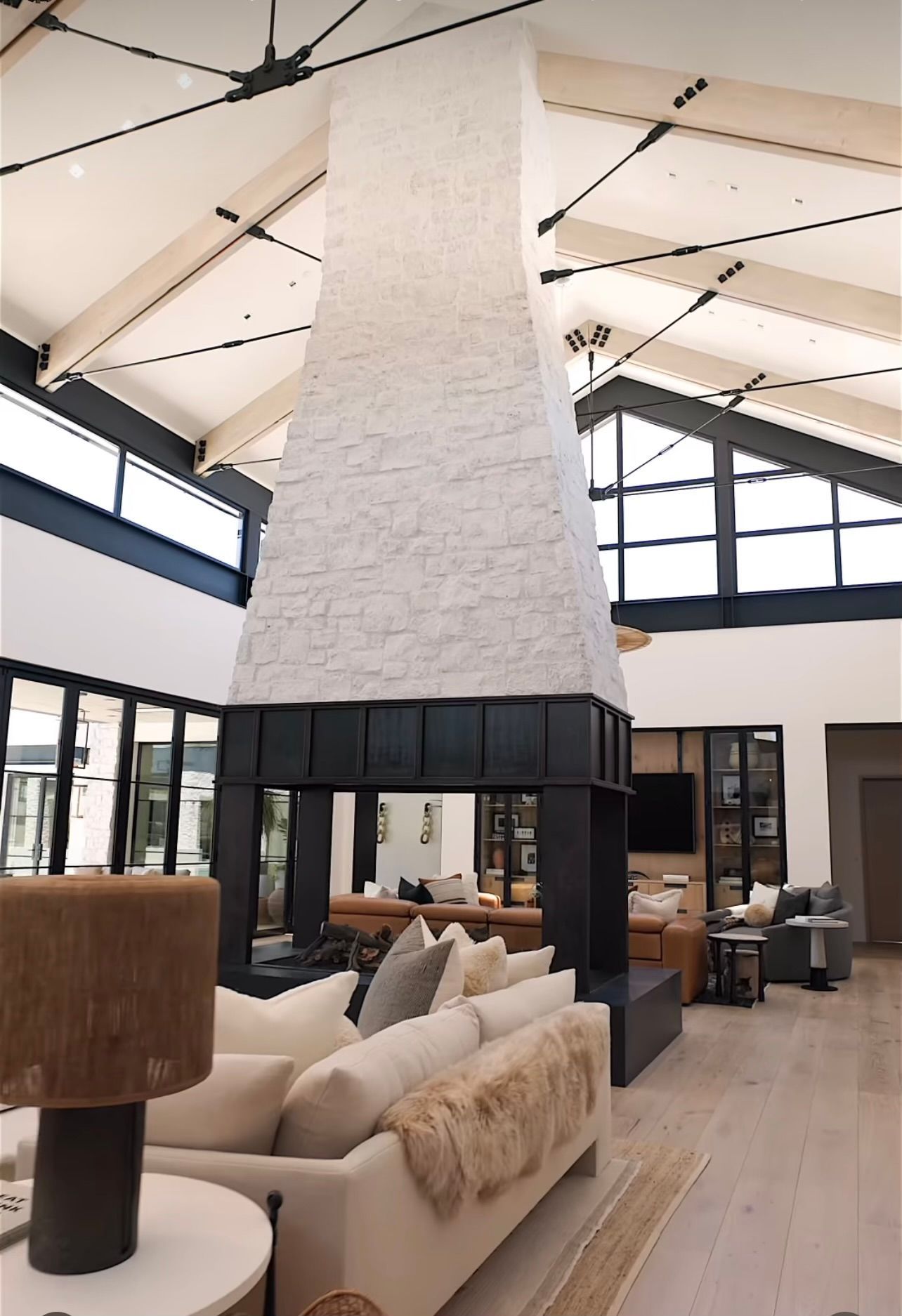 Modern lounge with a tall white chimney, beige sofas, wood floors, and large windows under a vaulted ceiling