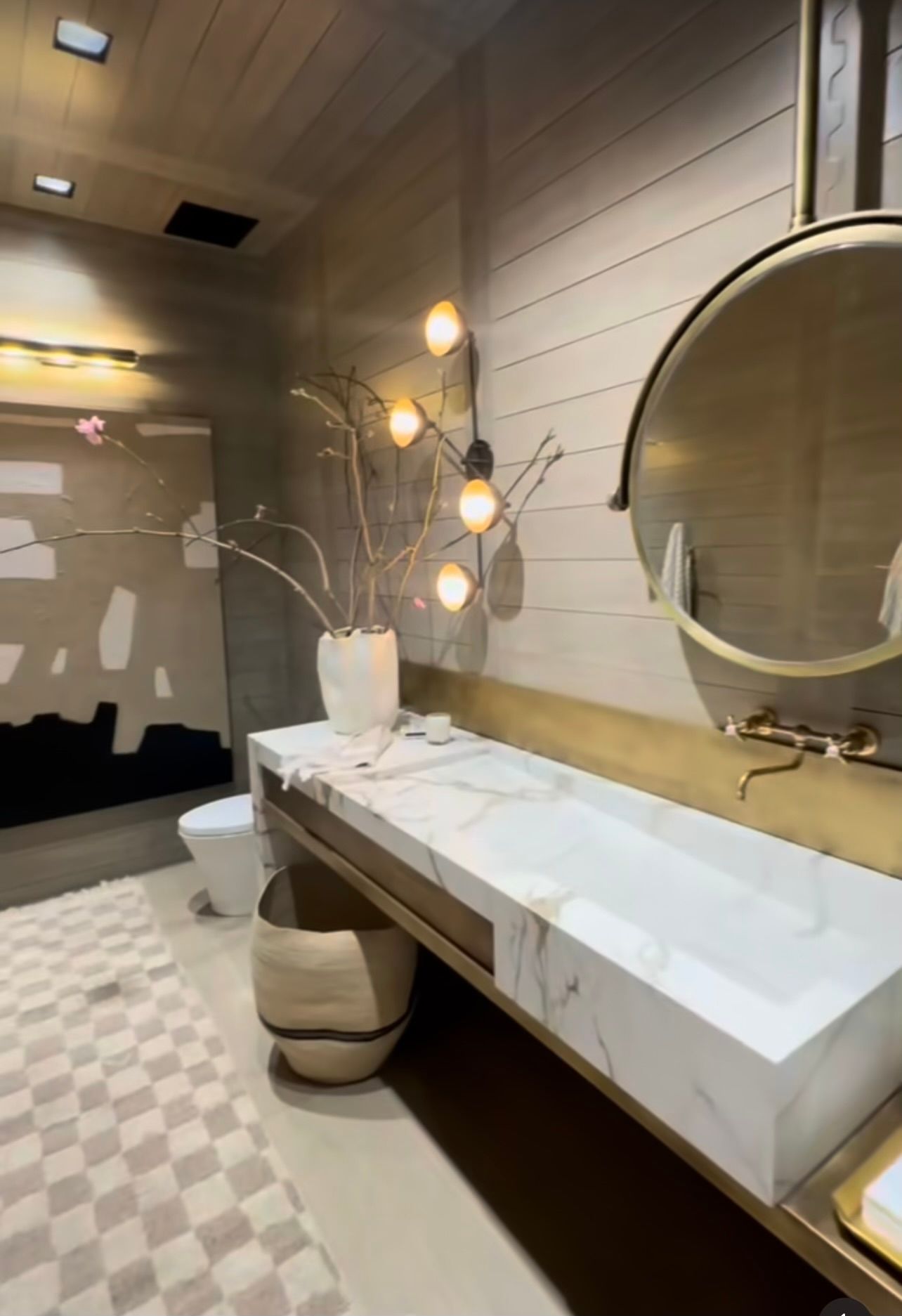 Modern bathroom with a double sink, round mirror, warm wall sconces, and checkered tile floor