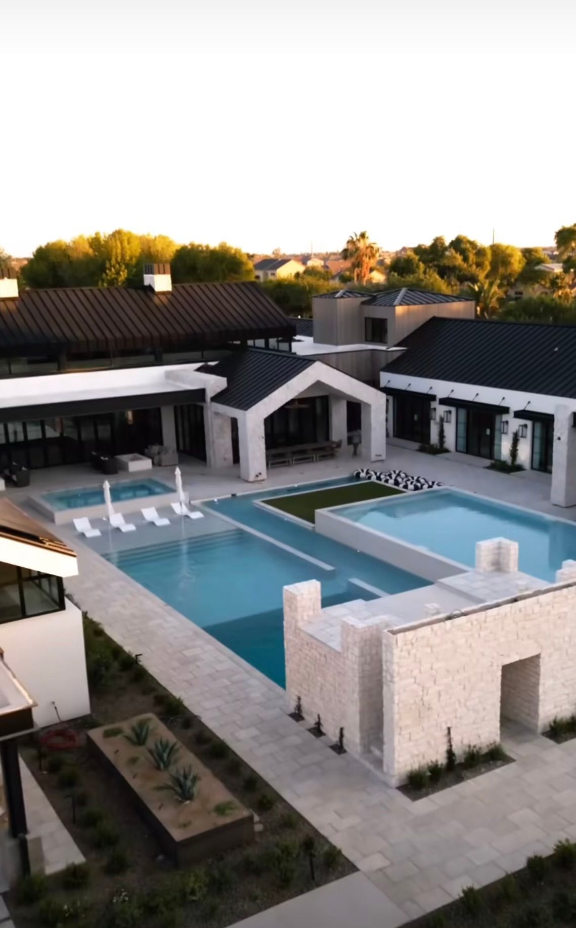 Modern courtyard with a pool, white lounge chairs, and dark-roofed buildings at sunset