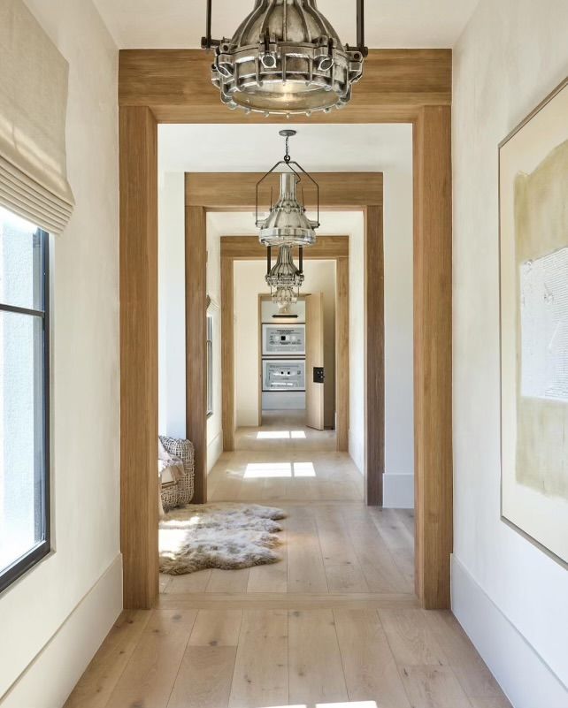 Bright hallway with wooden beams, hanging lanterns, pale walls, and light wood floors leading to a distant room