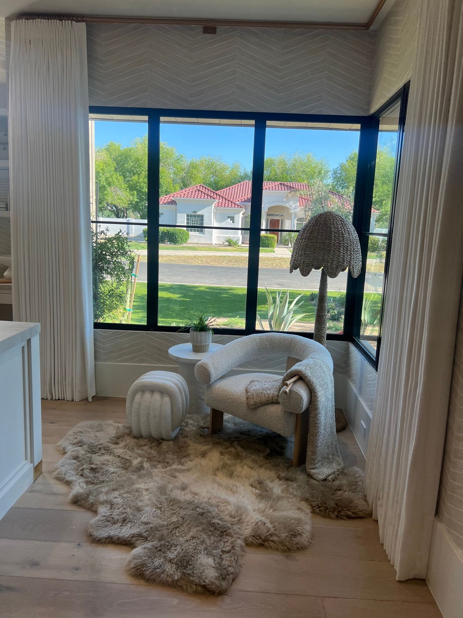 Cozy room with shaggy rug, patterned chair, white curtains, and large window view of a sunny neighborhood.