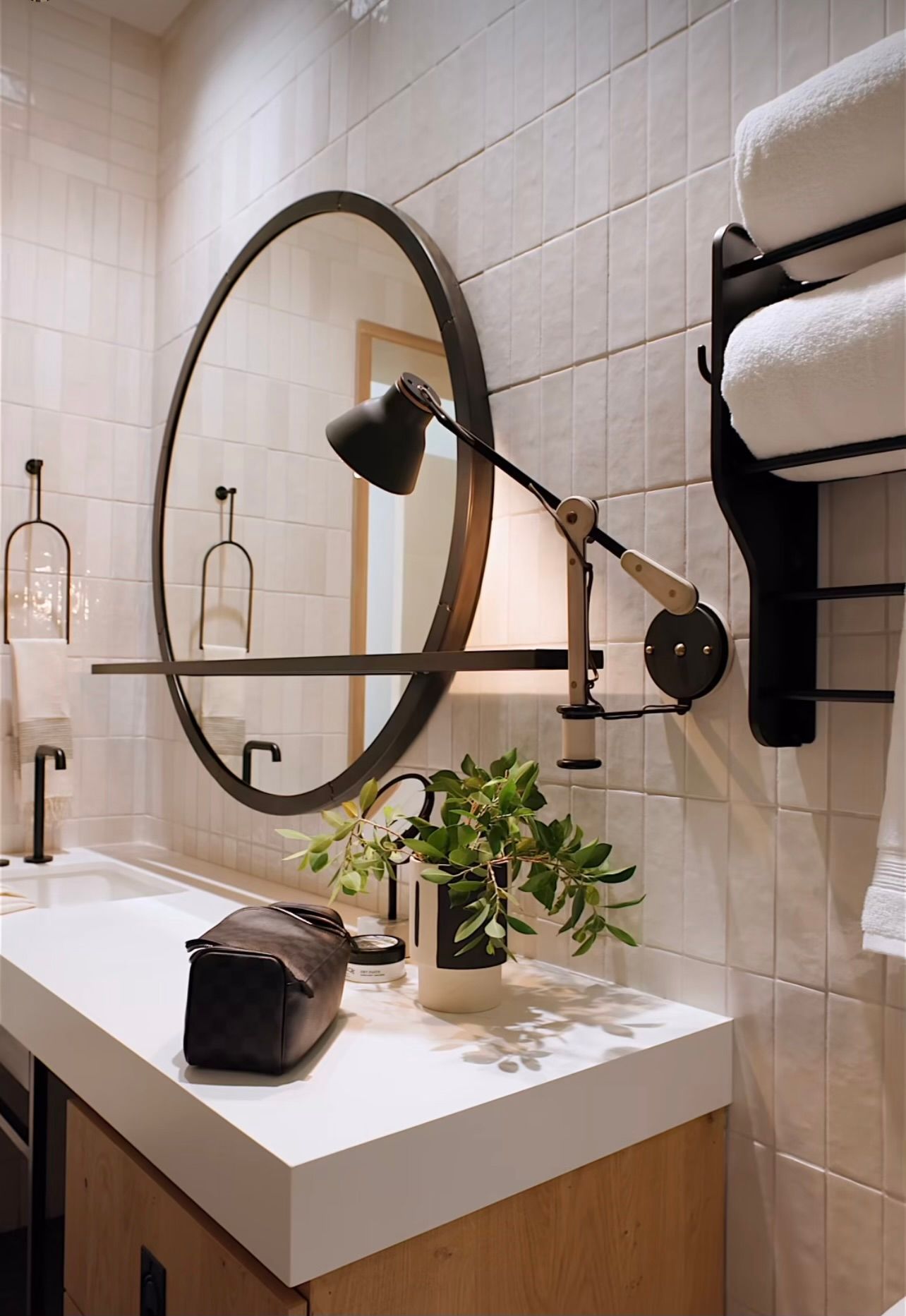 Modern bathroom vanity with round mirror, black sconce, towels, and plant on white countertop.