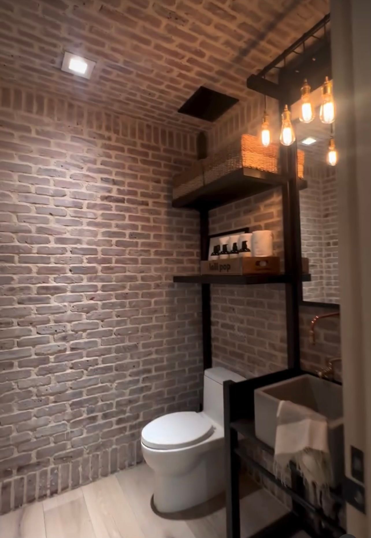 Modern bathroom with brick walls, hanging lights, toilet, and towel shelf beside a sink mirror