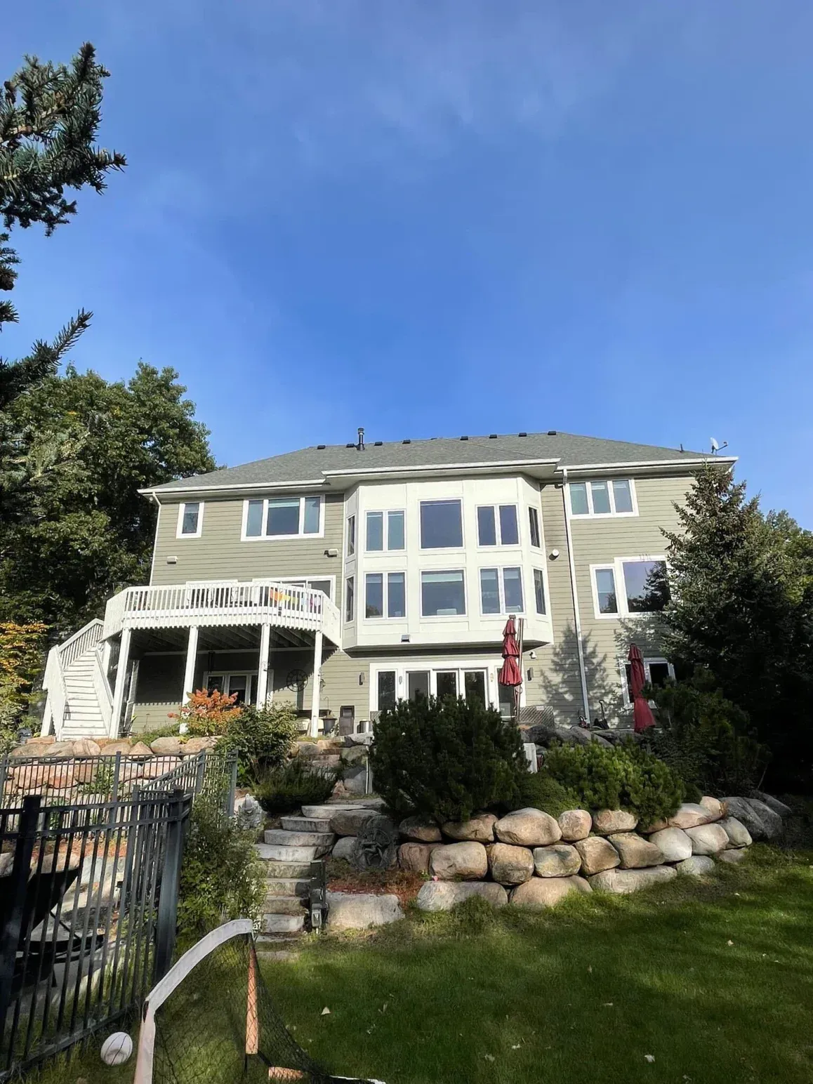 Back view of a large two-story house with green siding, surrounded by landscaping, under a blue sky.