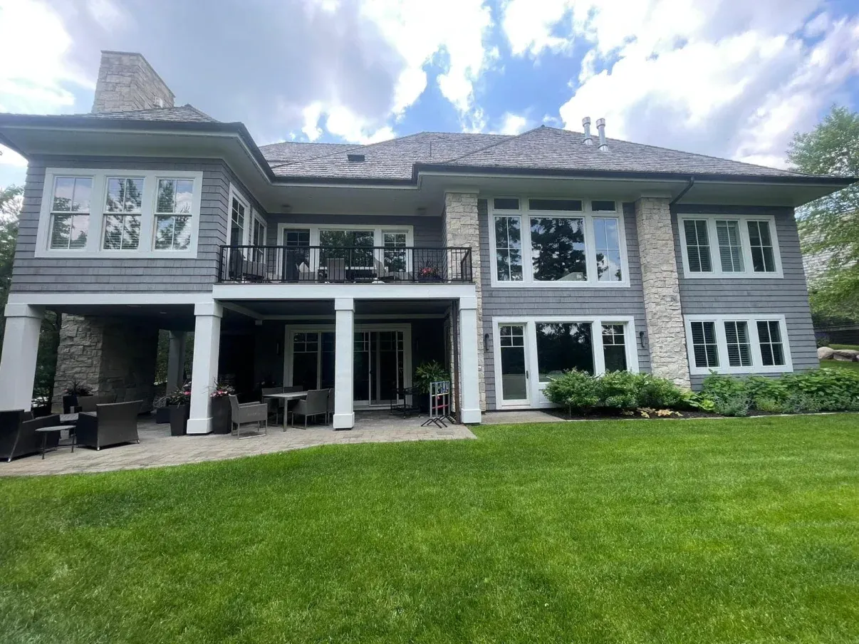 Back view of a large, gray and stone house with a balcony and patio, on a green lawn, under a cloudy sky.