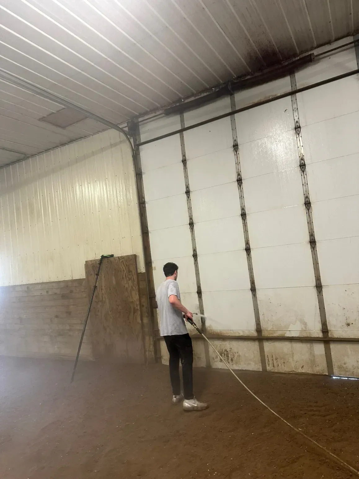 Person sprays water on a wall inside a building with a dirt floor.