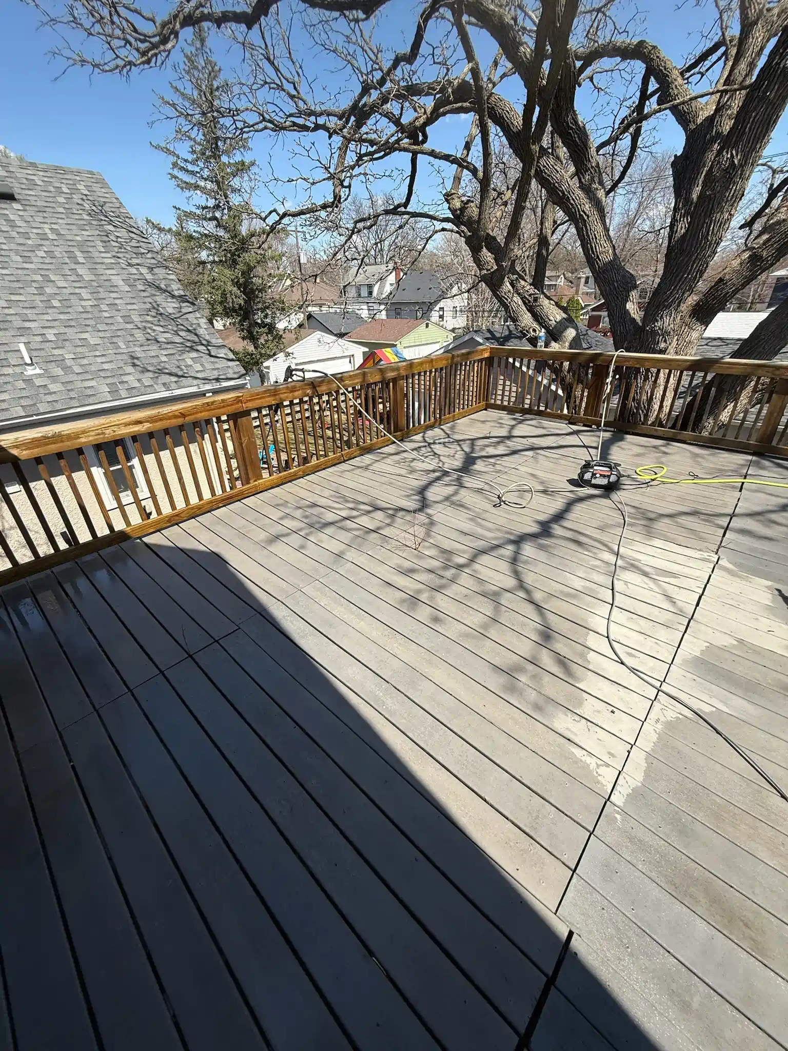 Wooden deck with railings, tree branches overhead, and a view of rooftops on a sunny day.