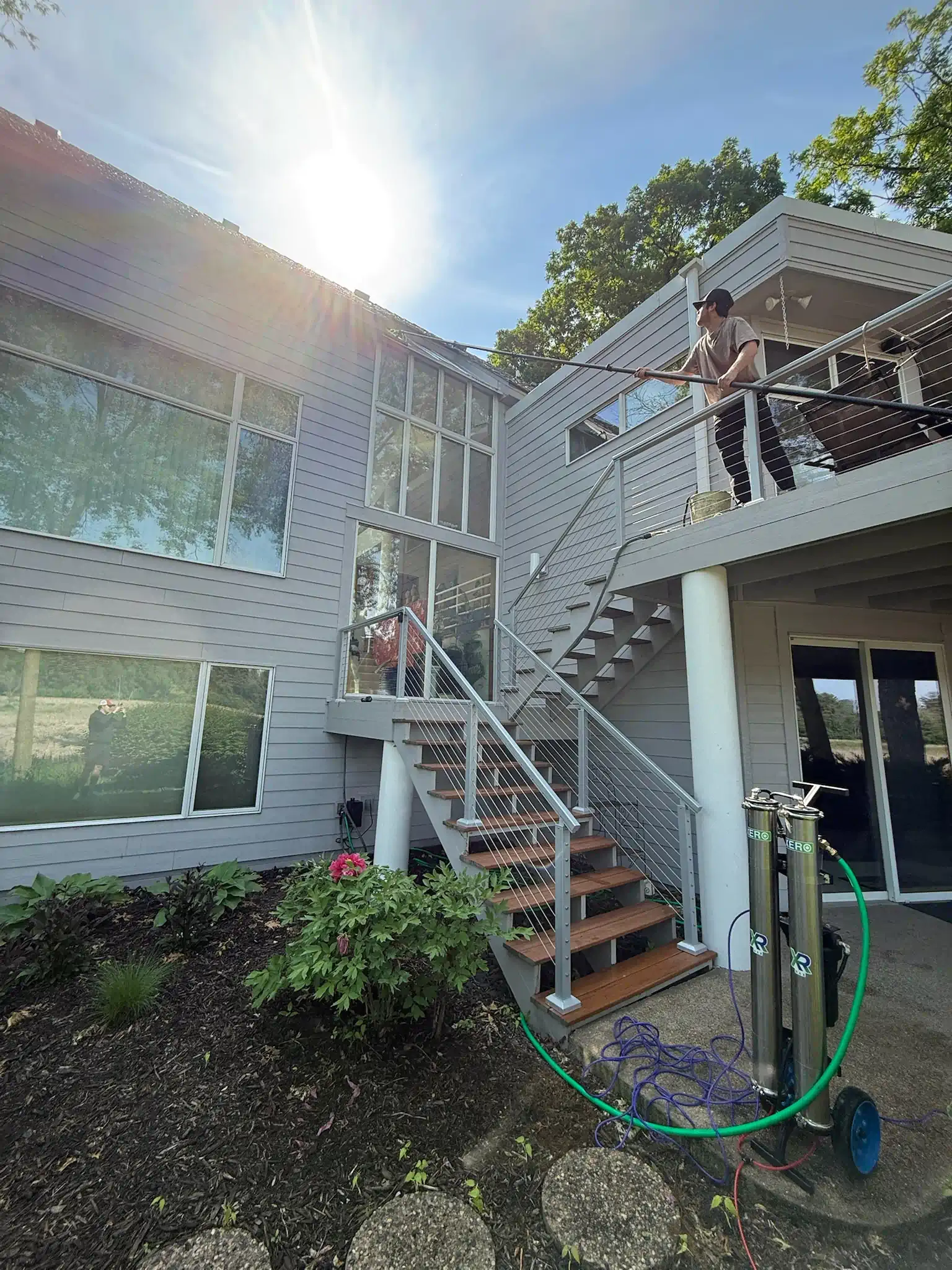 A person washing windows on a two-story beige house with a water-fed pole. Green lawn with bushes and trees.