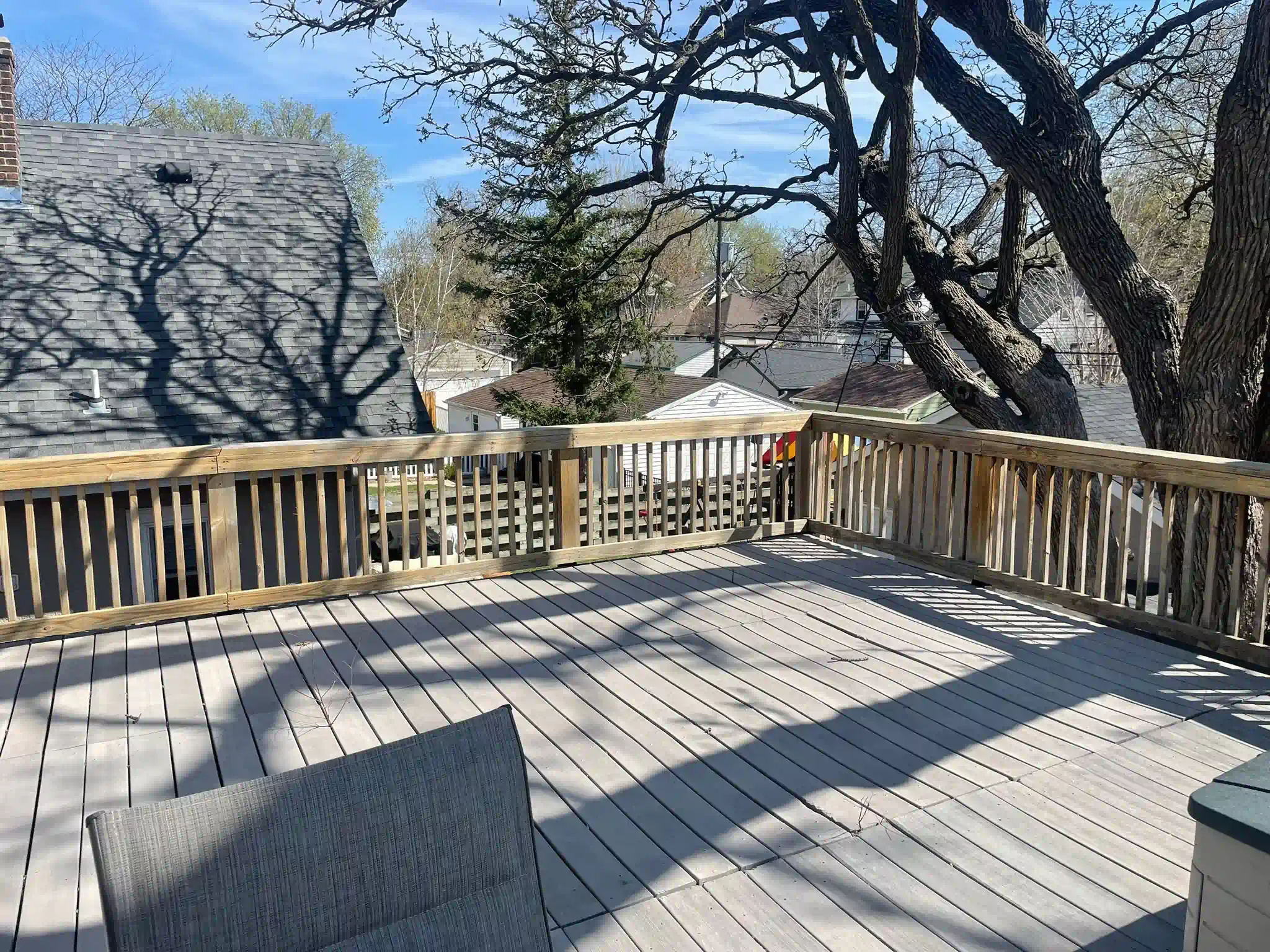 Wooden deck with gray planks and railing, shadowed by tree branches on a sunny day.