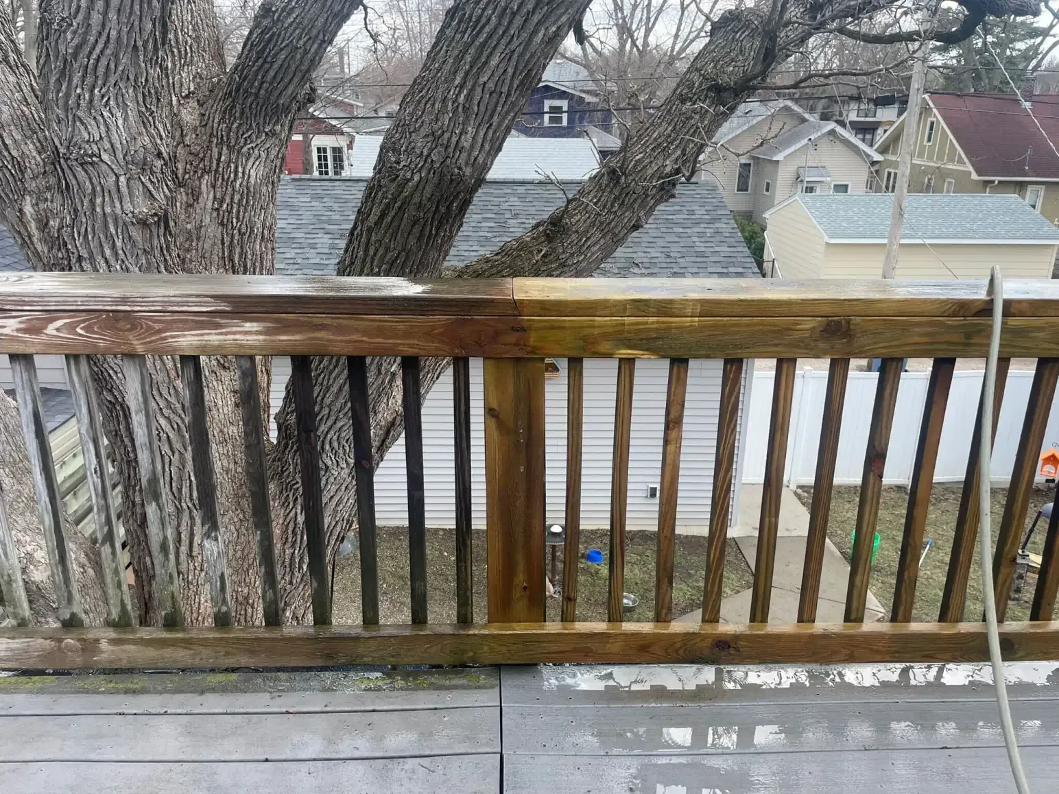 Half-cleaned wooden deck railing, showing the difference before and after washing. A tree and houses are in the background.