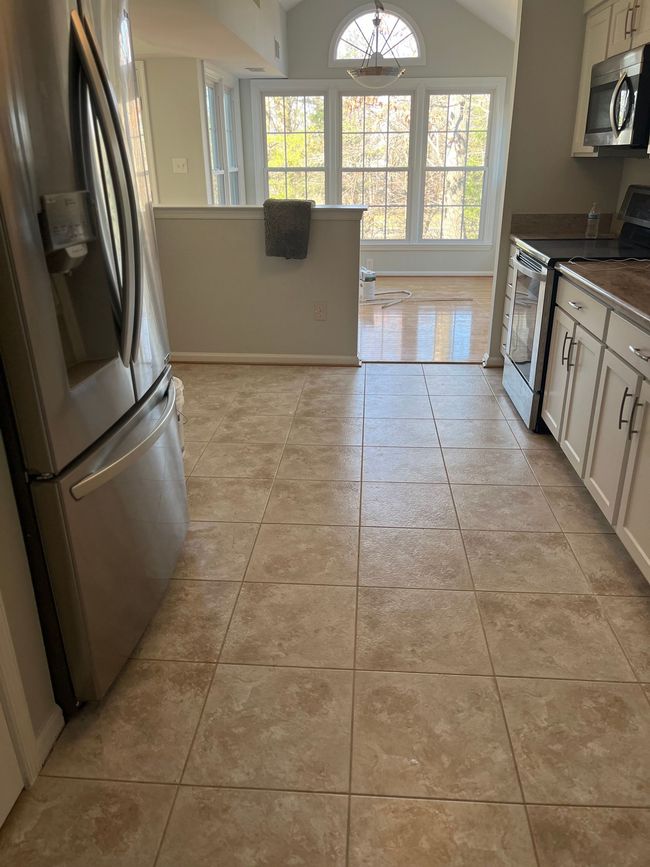 Kitchen with beige tile floor, stainless steel refrigerator, and white cabinets. Sunny window in background.