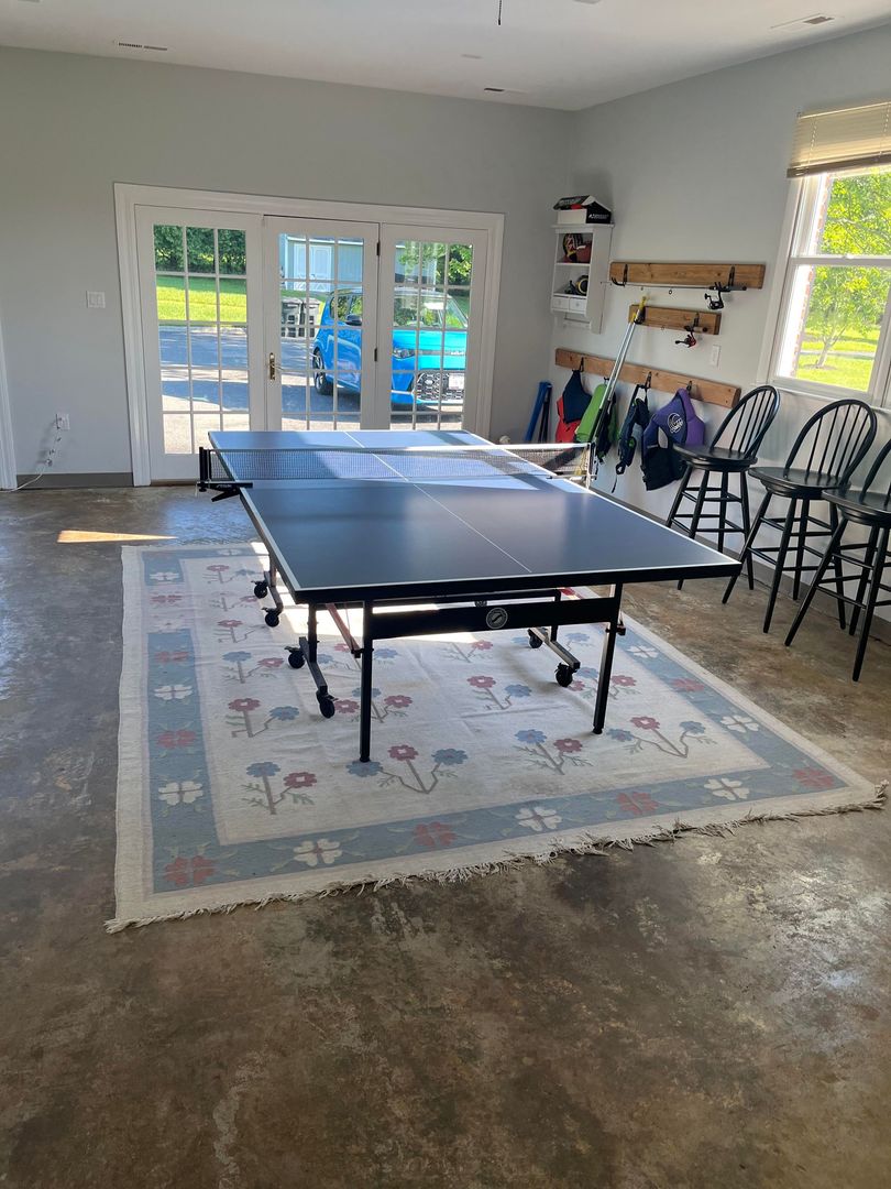 Ping pong table in a garage, set on a rug. Doors open to a blue car. Bar stools line the side.