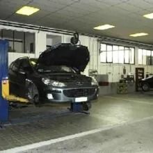 Auto sollevata in officina. Cofano aperto, meccanico al lavoro. Interni, luci e attrezzi visibili.