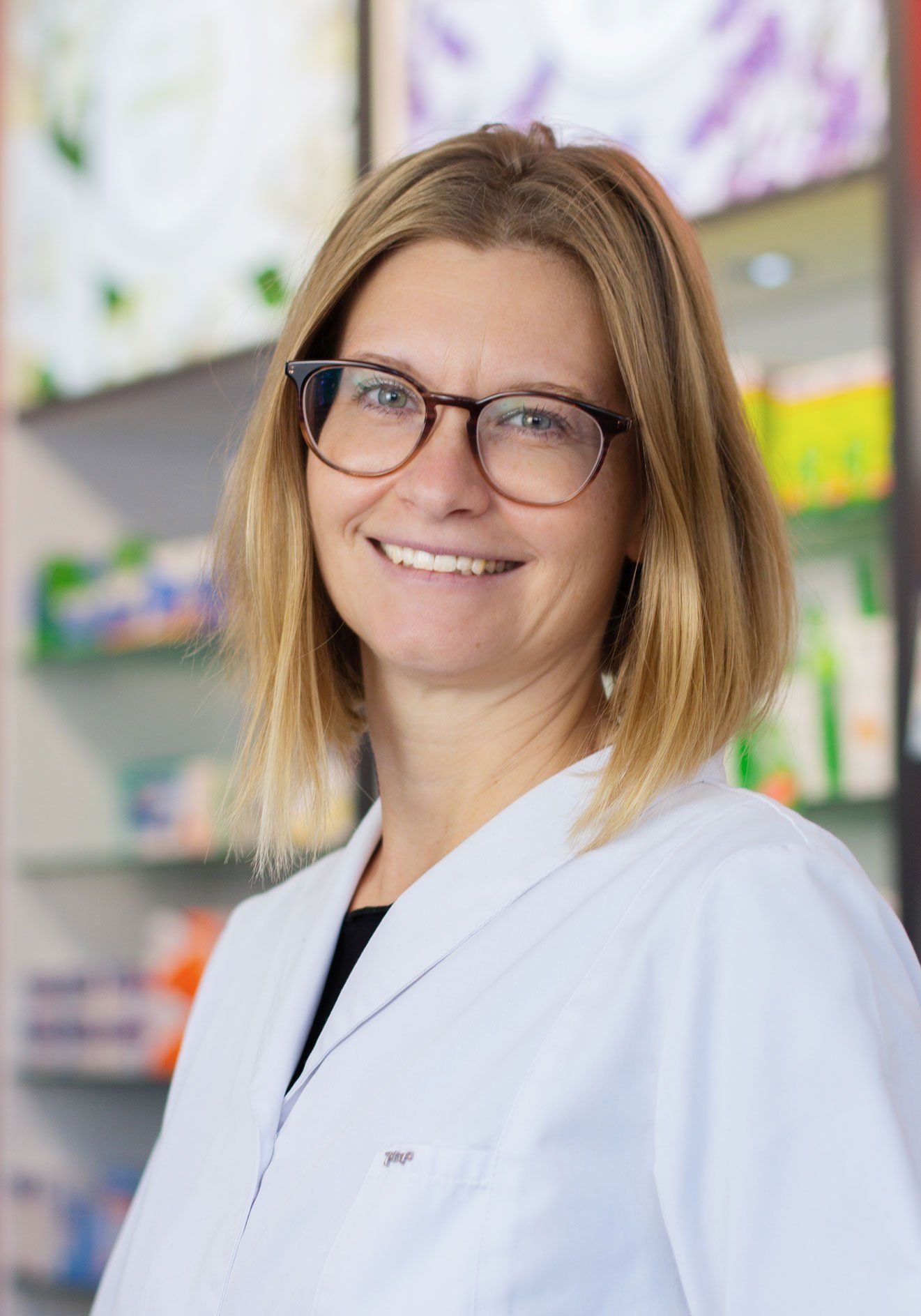 Eine Frau mit Brille und weißem Kittel lächelt in einer Apotheke.