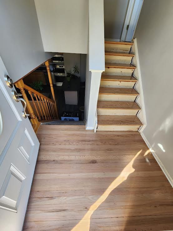 Entryway with light wood floor leading to a staircase. The staircase has bare steps and a wooden railing.