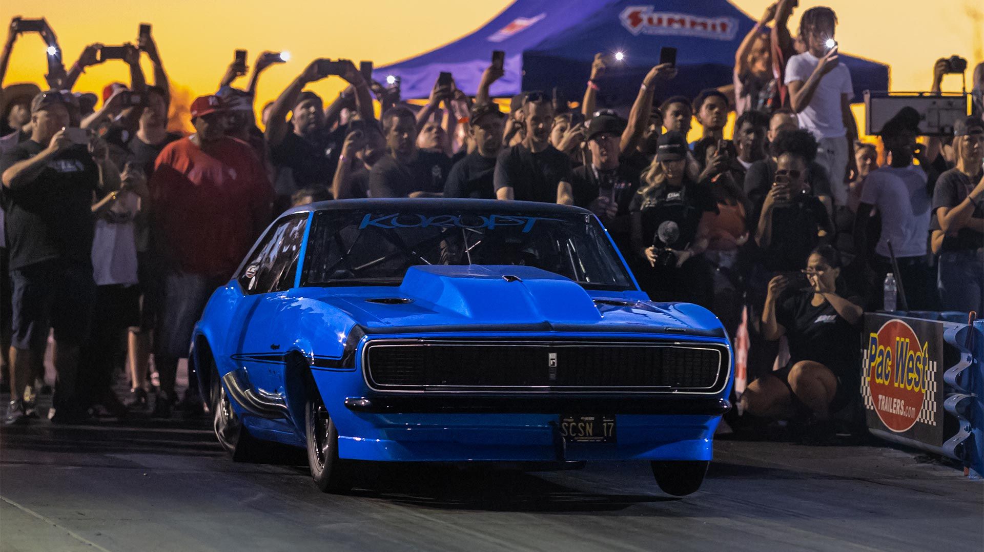 Blue race car on track, crowd cheering with phones, sunset sky.