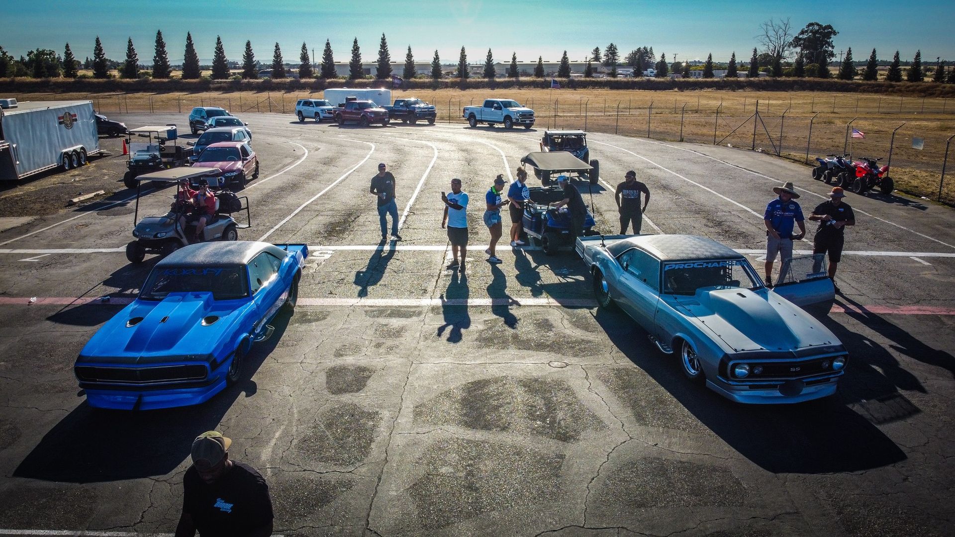 Two drag racing cars lined up on a track, people standing nearby.