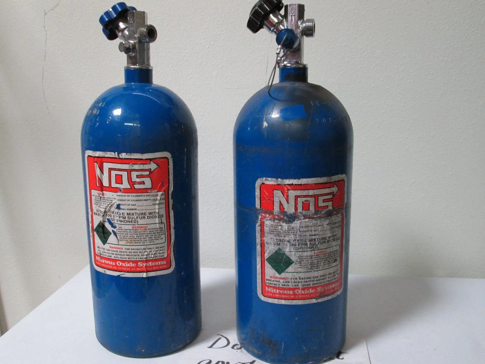 Two blue NOS nitrous oxide cylinders on a white surface, with red and white labels and silver valves.