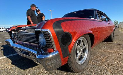 Classic orange car with racing stripe, two men nearby on asphalt under a blue sky.