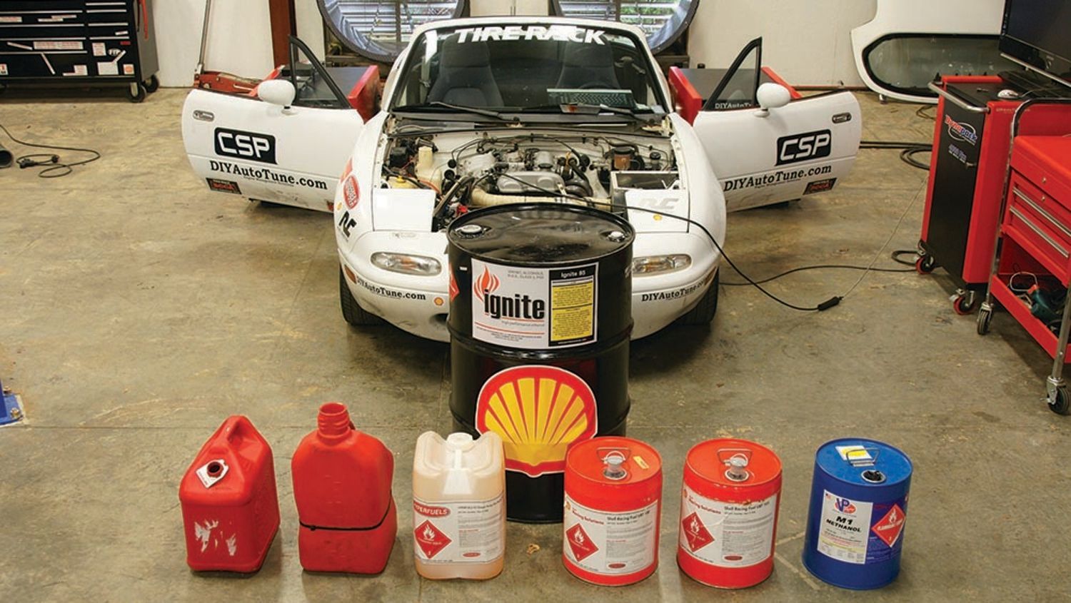 White car with open doors and engine bay open, in front of a variety of fuel containers in a garage setting.