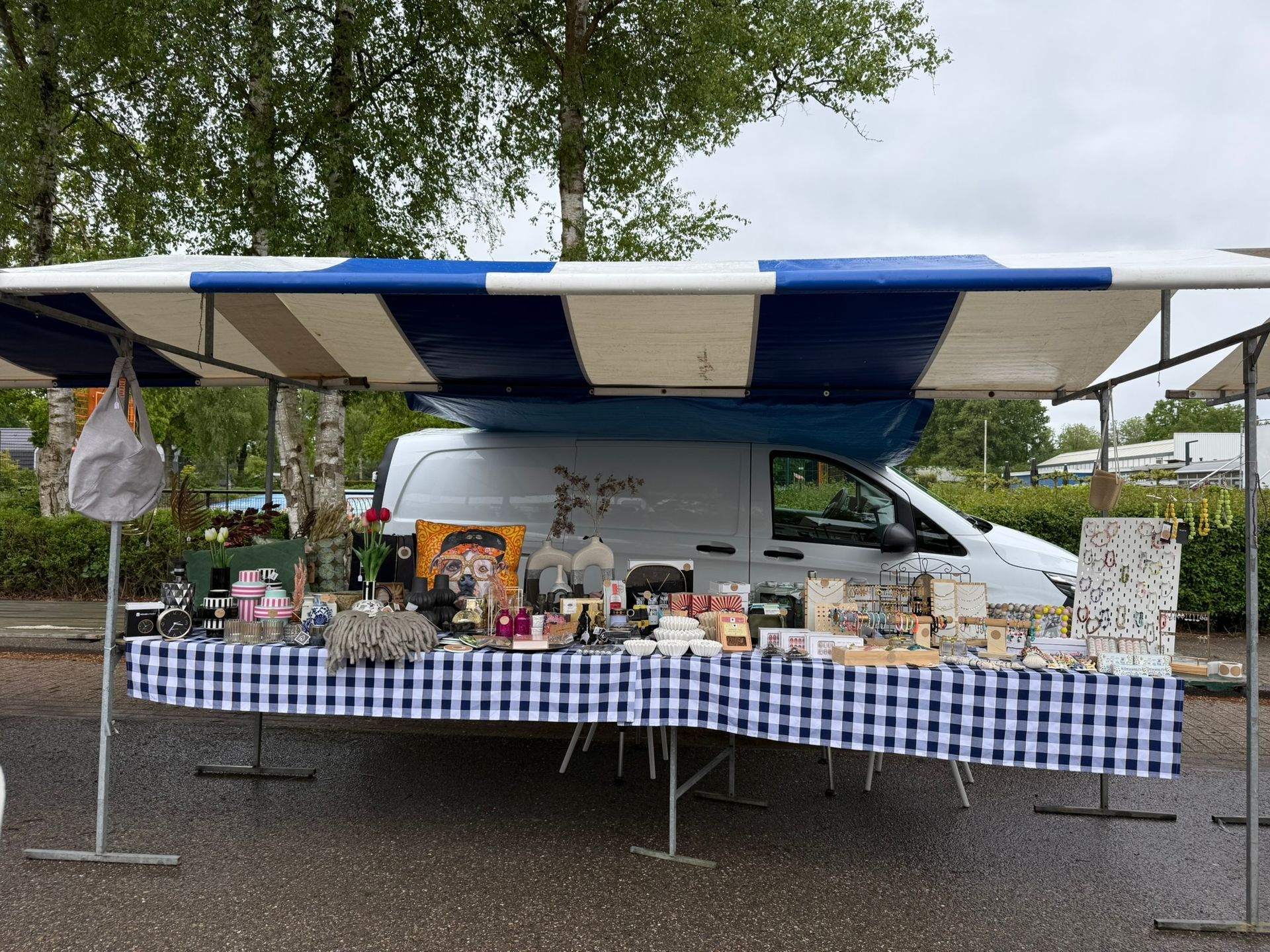 Marktkraam in de openlucht met een blauw-wit afdak, een tafel vol koopwaar en een witte bestelwagen op de achtergrond.