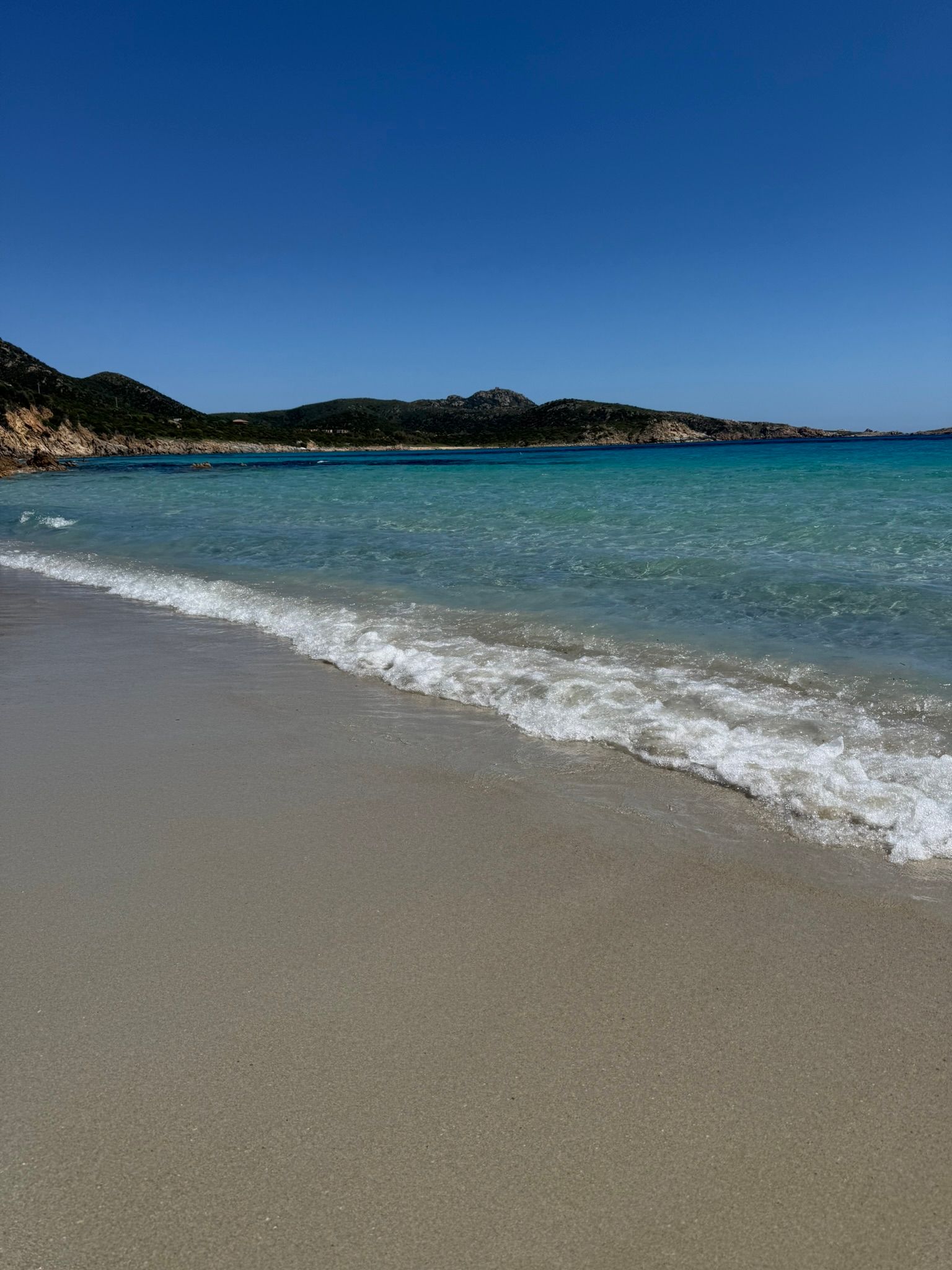 Zonnig strand met helderblauw water, zachte golven en in de verte een rotsachtige kustlijn van Sardinië.