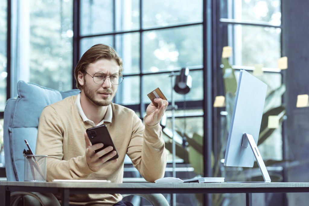 A confused-looking man is holding his mobile phone on one hand and his credit card on the other.