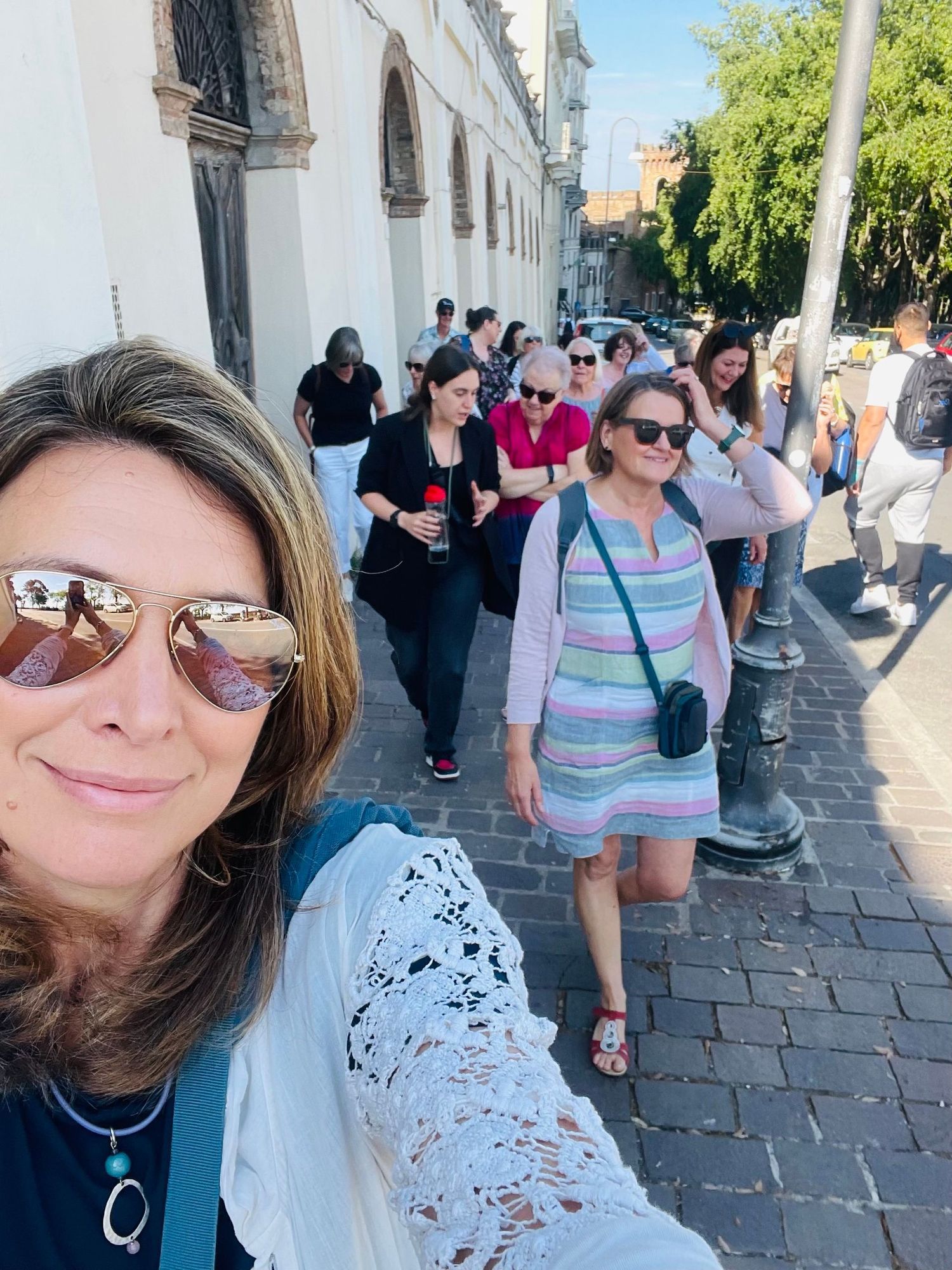 People walking along a sunny cobblestone street beside a white building, with a woman taking a selfie in front.