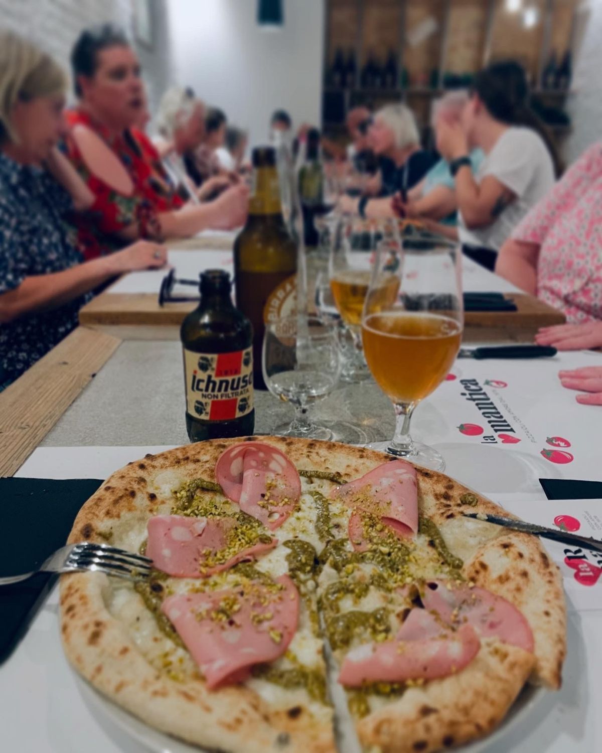 Table with pizza and drinks at a lively restaurant gathering; people sit chatting in the background.