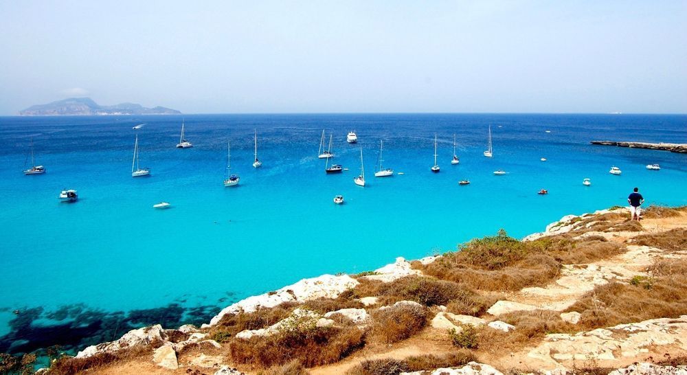 Turquoise bay with sailboats anchored offshore beside a rocky shoreline under a clear blue sky