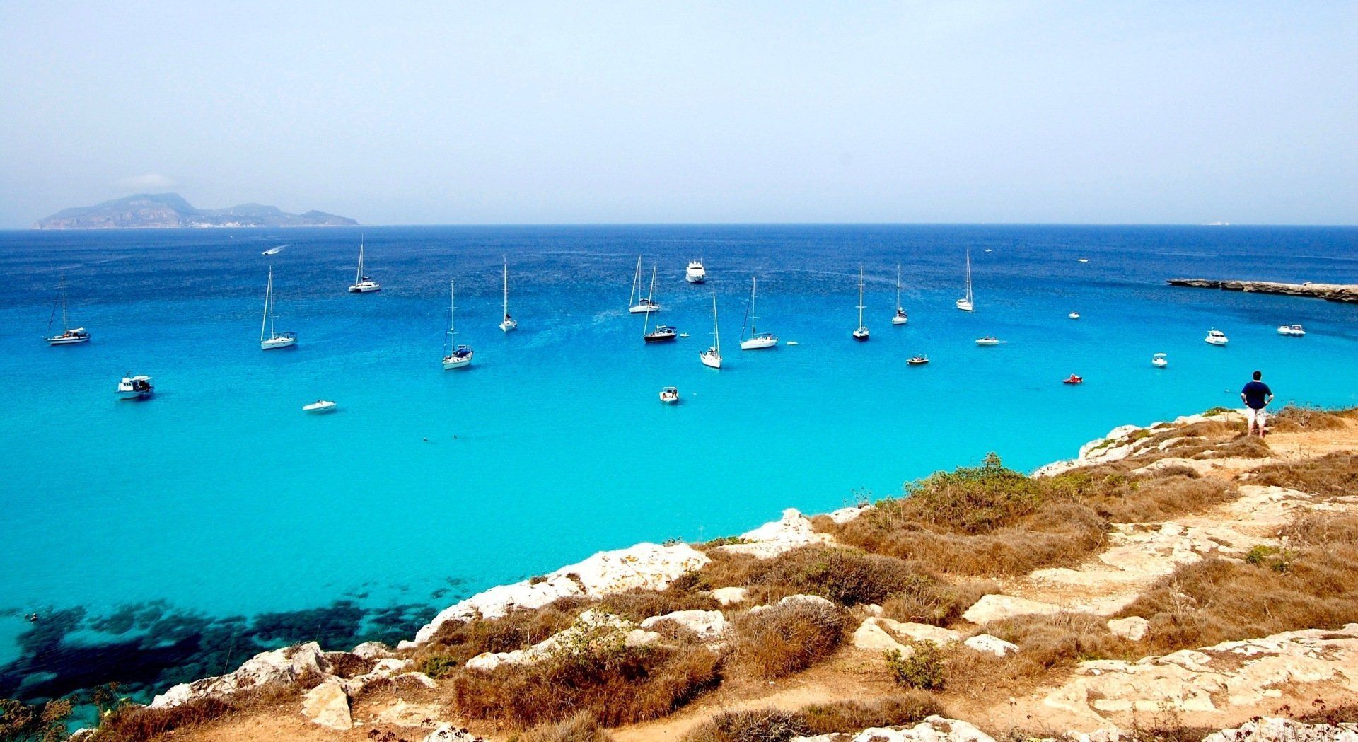 Turquoise sea with anchored sailboats and a rocky shoreline under a clear blue sky