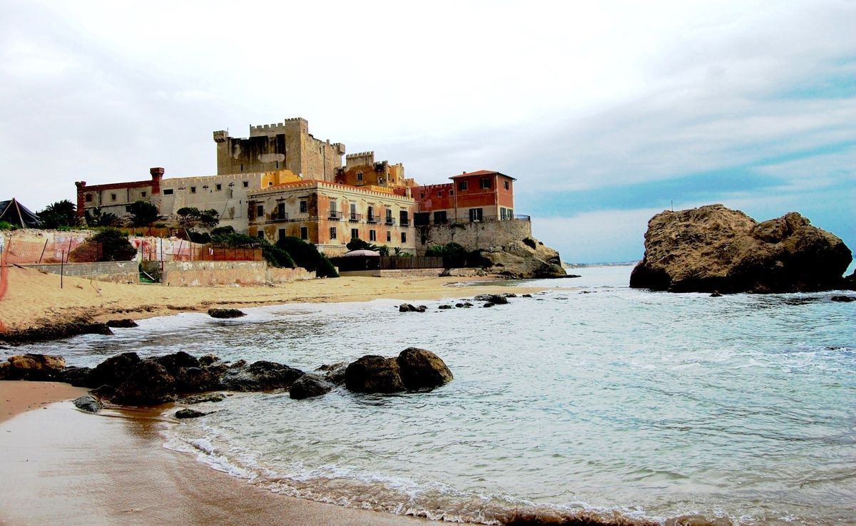 Seaside village with stone buildings on a rocky shore beside calm blue water under a cloudy sky
