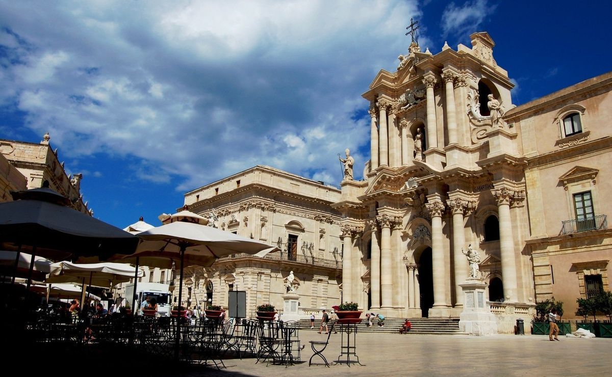 Historic cathedral plaza with ornate stone facade, café tables, and a bright blue sky