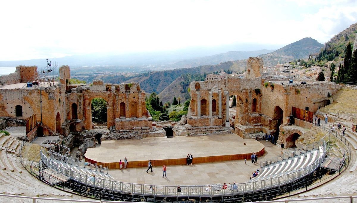 Ancient open-air amphitheater with stone seats, arched stage, and mountain backdrop under clear sky