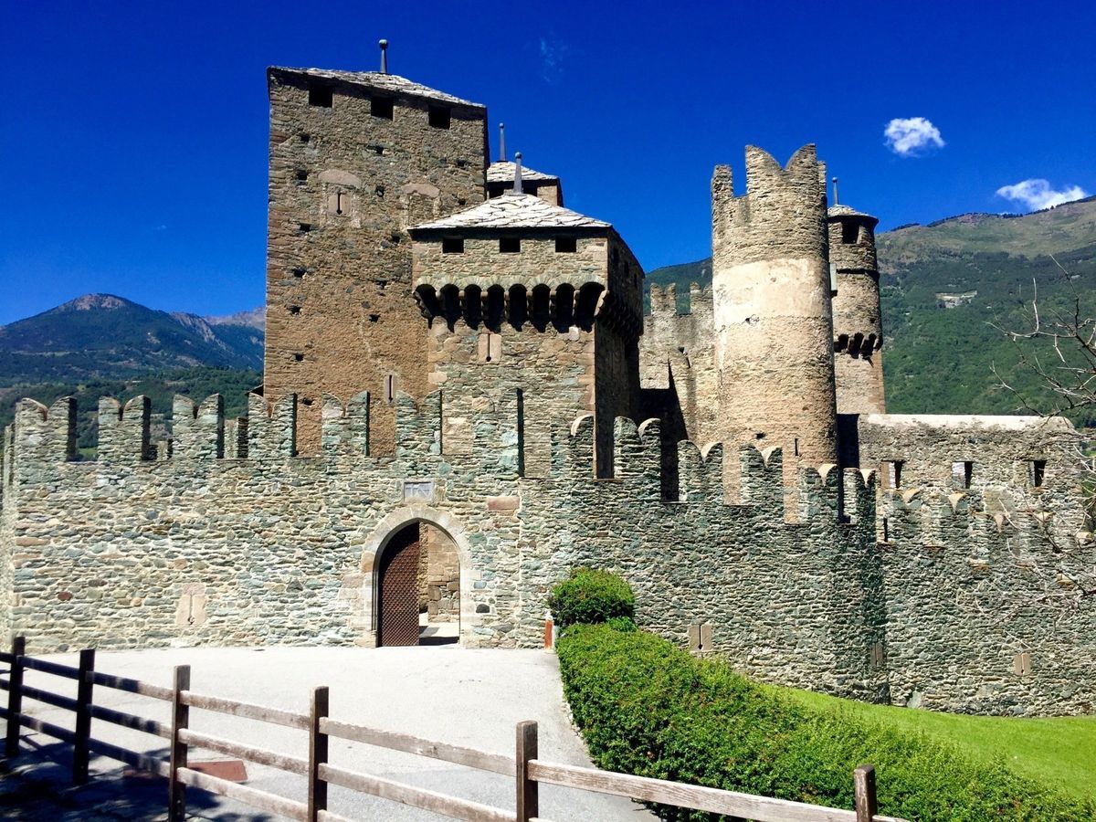 Stone castle with towers and battlements behind a fortified wall, set against mountains and a blue sky