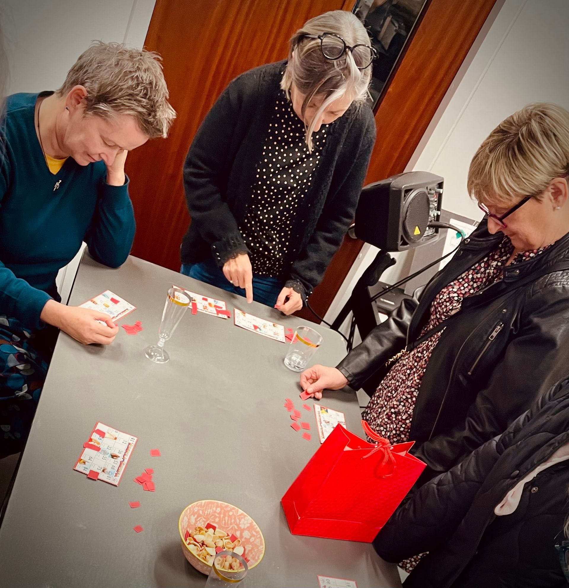 Three people play cards at a table with a bowl of candies and a red gift bag.