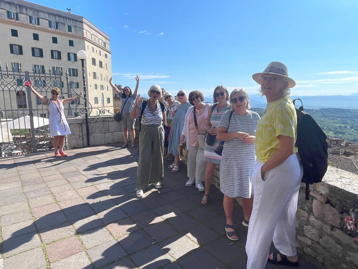 Group of tourists standing on a stone terrace by a historic building, with a scenic valley view under a blue sky.