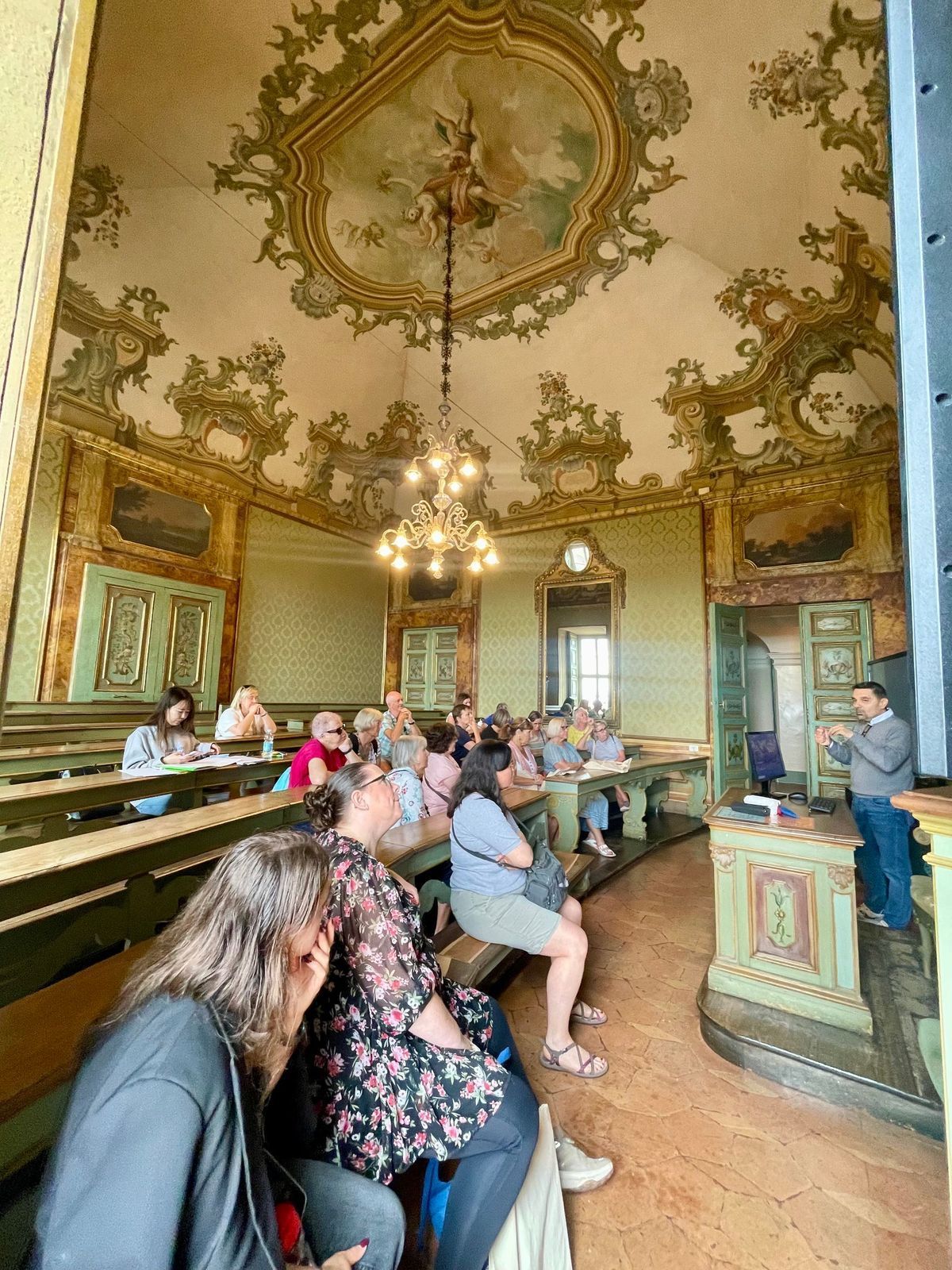 Tourists seated in an ornate historic hall with gilded ceiling, chandeliers, and painted walls