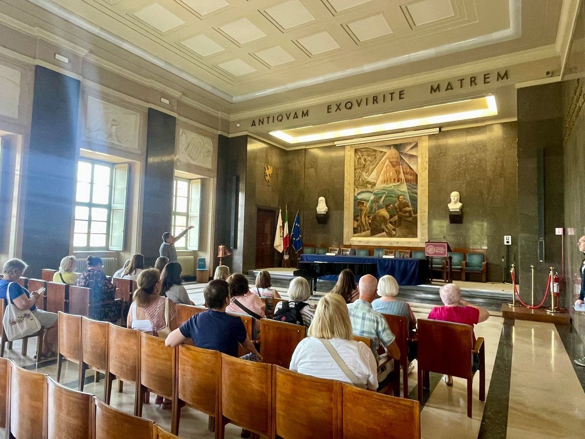 People seated in a formal room facing a podium and portraits, with a large banner above.