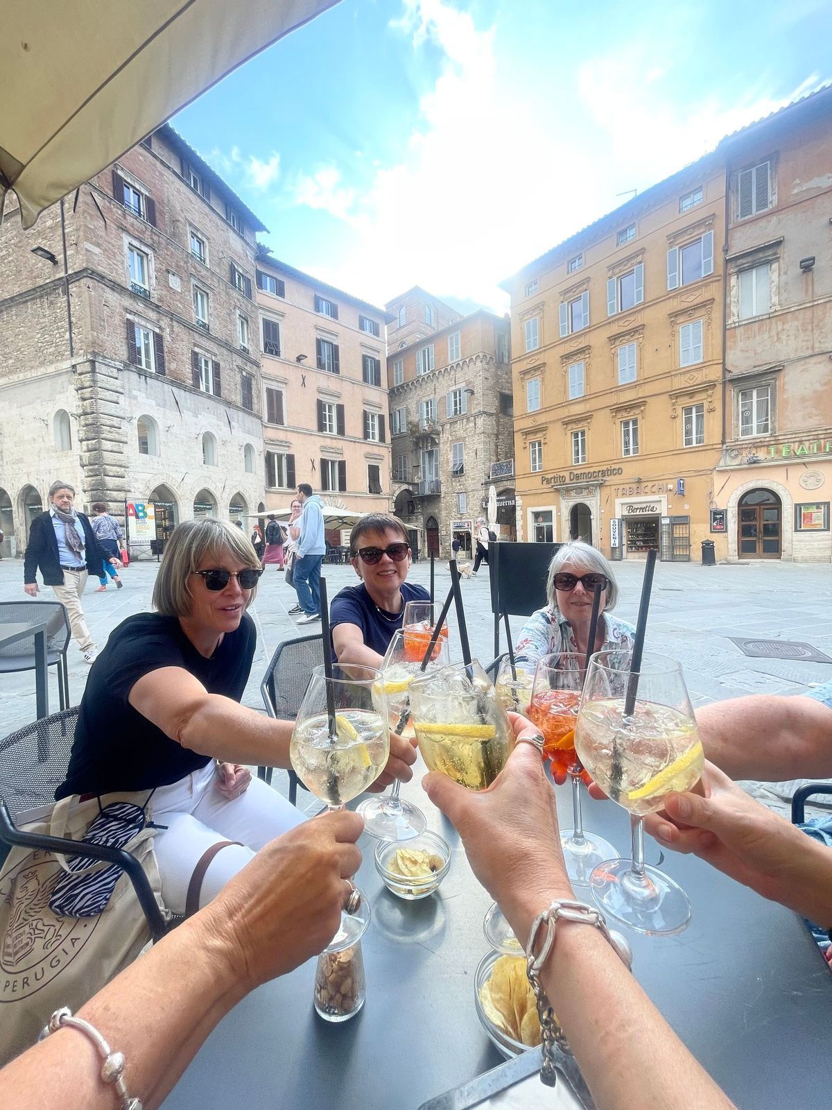 Friends clink wine glasses at an outdoor café in a sunny European square.
