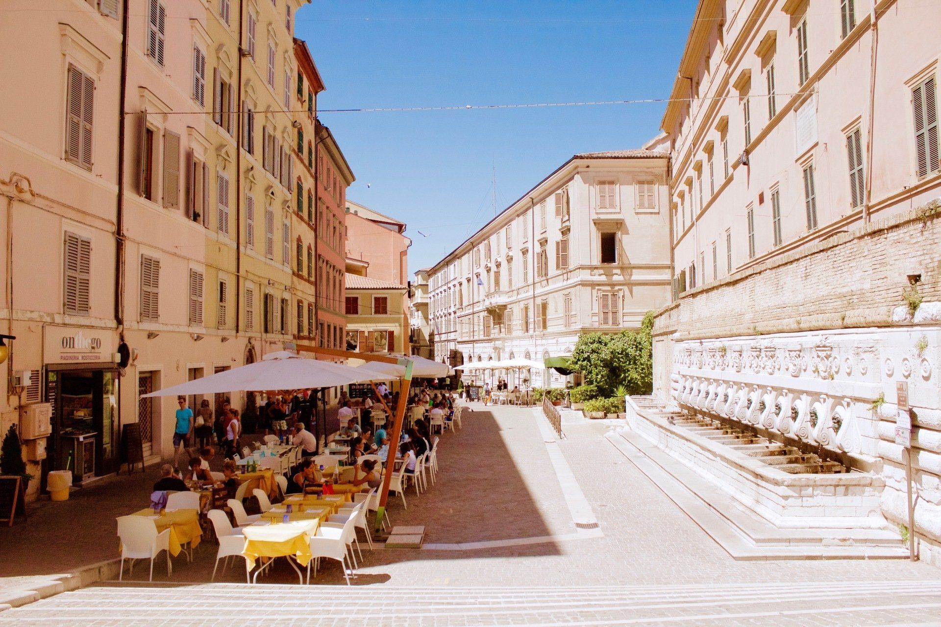 Sunny European street with outdoor café seating beside historic buildings and a stone wall in a narrow alley