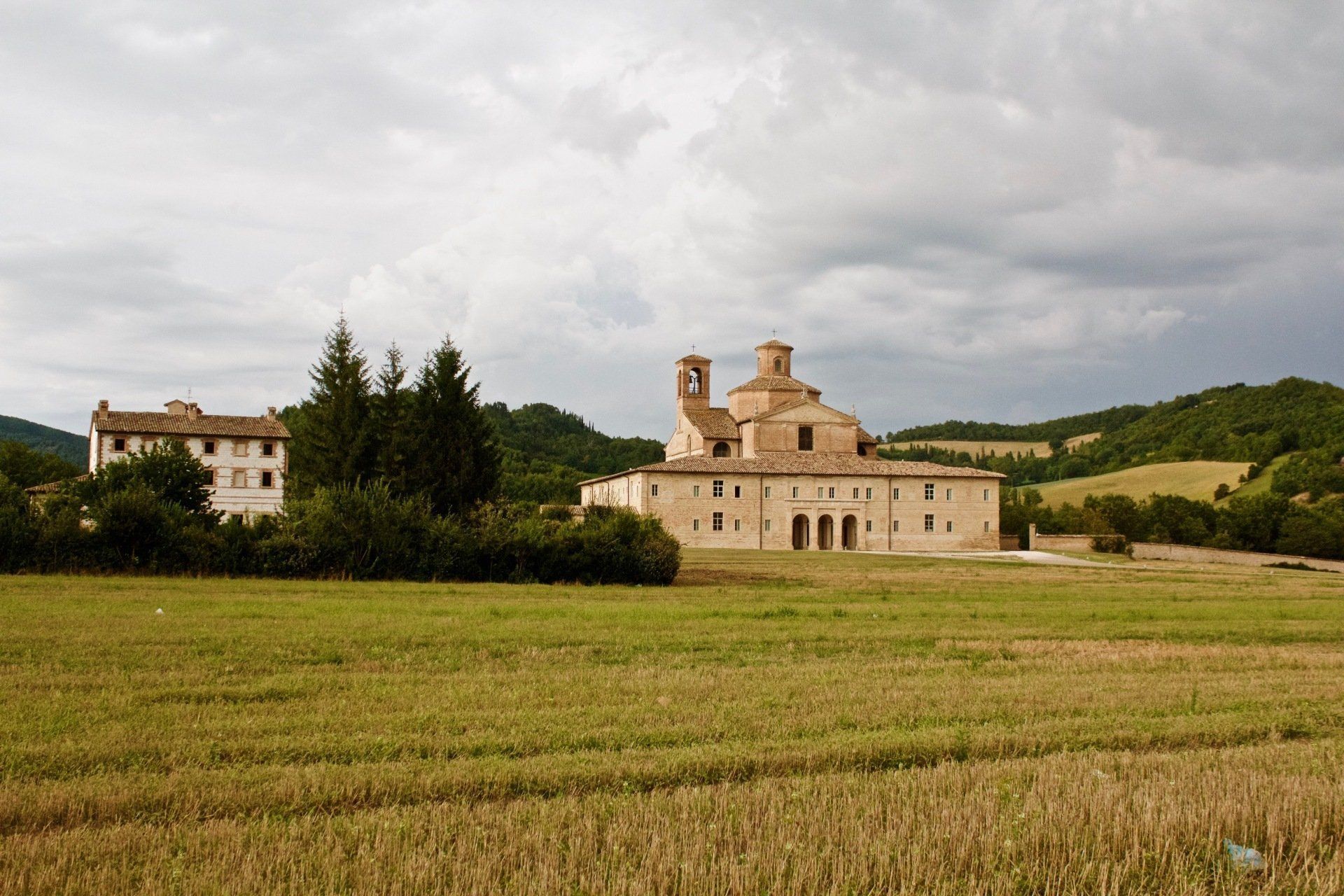 Rural monastery with a stone church and buildings beyond a green field under cloudy skies