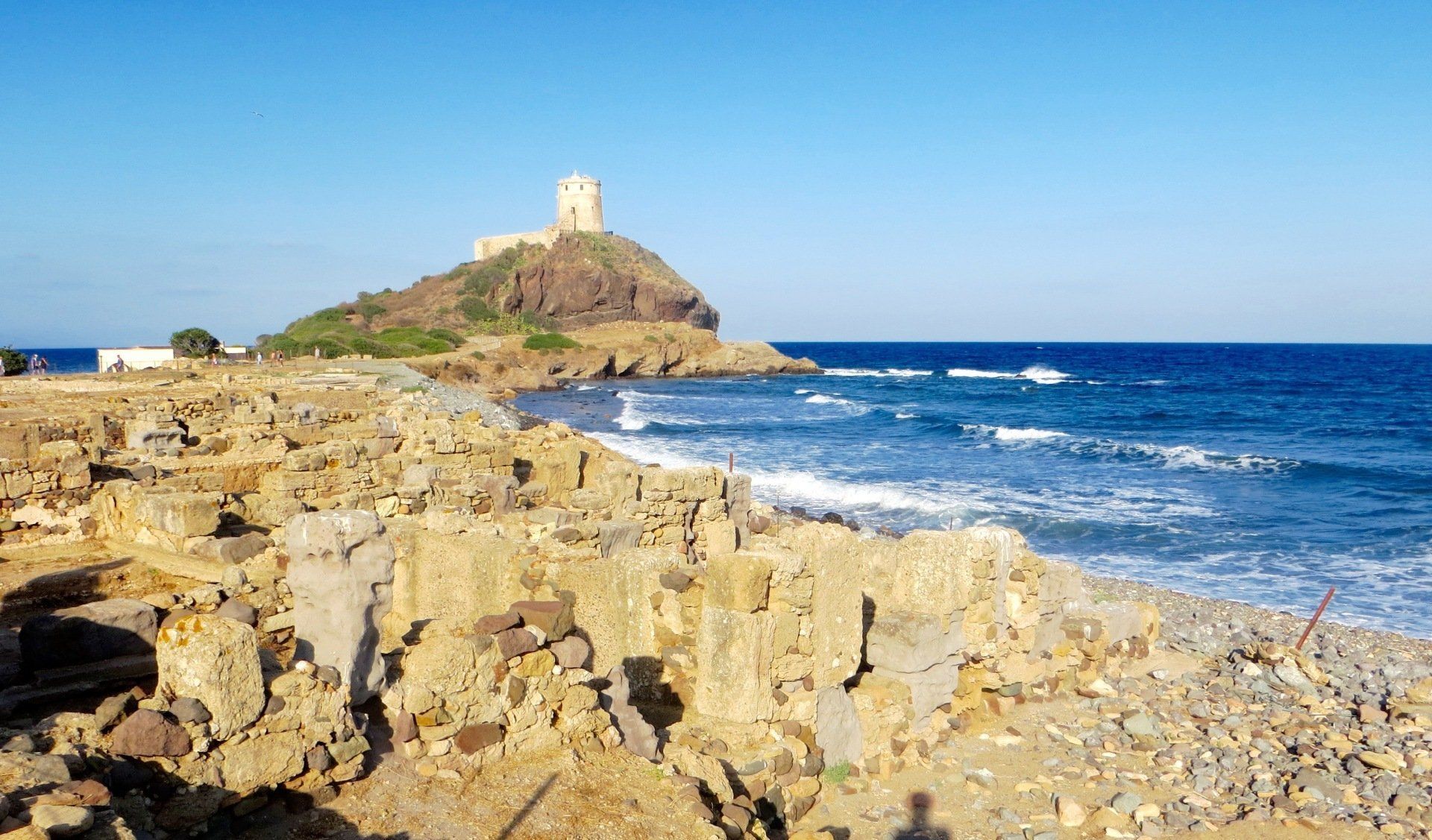 Rocky seaside coastline with blue waves and a small white tower on a hilltop in the distance