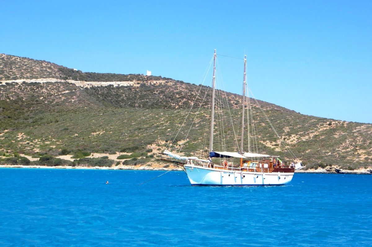 White sailboat on bright blue water near a rocky green coastline under a clear sky