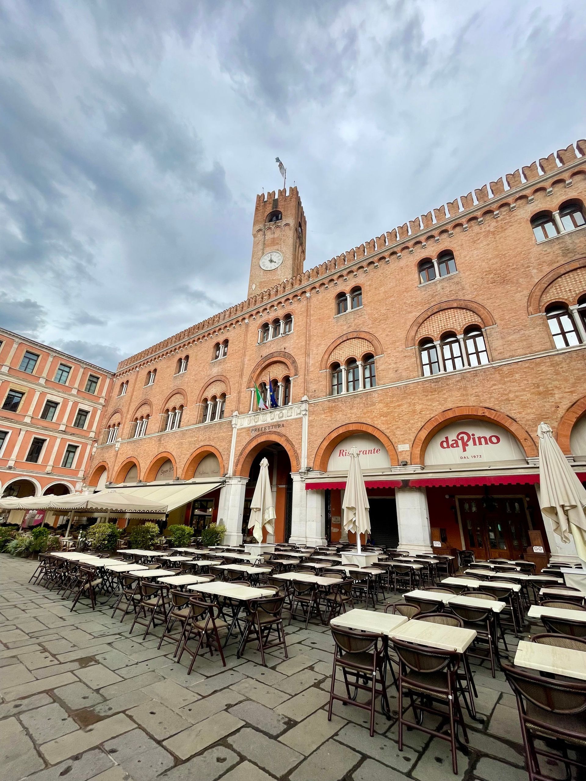 Historic brick building with tower and arched windows above a café terrace with outdoor tables