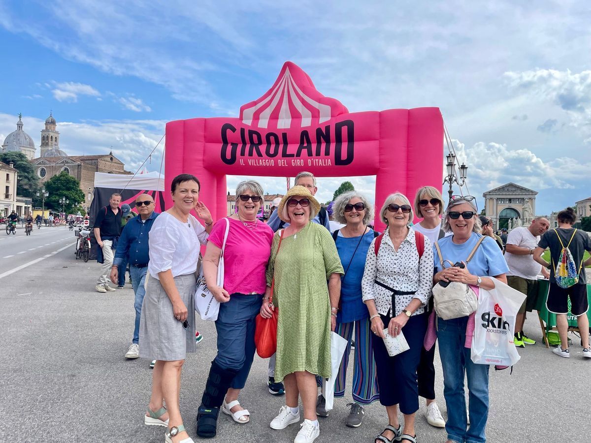 Group posing before a pink CIGROLAND arch on a cloudy street, with pink-clad performers and spectators.