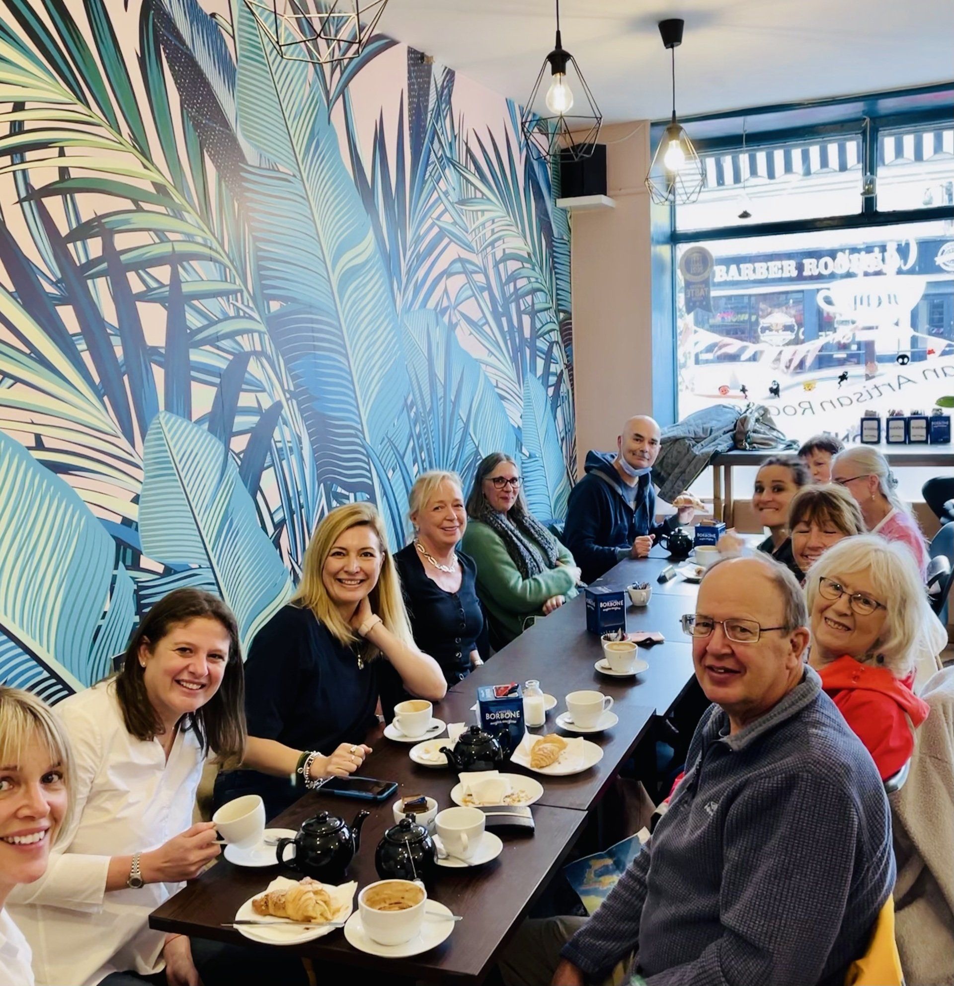 Group of people smiling at a café table beside a colorful leaf mural, with coffee and pastries