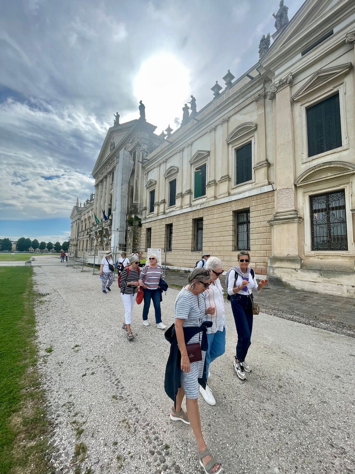 Tour group walking beside a grand historic building on a gravel path under a cloudy sky